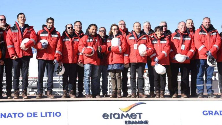 Un grupo de personas con chaquetas rojas y botas de seguridad, posando frente a una plataforma que lleva el logo de "Eramet Eramine". Algunos sostienen cascos de protección. Detr?s del grupo se ven estructuras que parecen ser parte de una planta industrial en un entorno de cielo despejado.