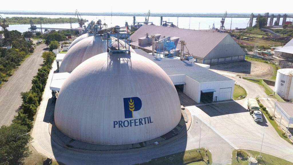 La imagen muestra en primer plano dos grandes estructuras de almacenamiento con forma de c?pula, con el logo de la empresa en la parte frontal. Al fondo, se puede observar parte del complejo industrial, que incluye otras edificaciones y estructuras relacionadas con el proceso de producción. Tambi?n se aprecian grúas y equipos de carga cerca de una zona portuaria.