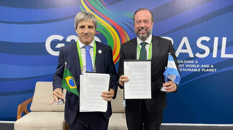 Luis Caputo y Alexandre Silveira, ministros de Argentina y Brasil, respectivamente, muestran a c?mara copias del acuerdo mientras sostienen banderas de ambos pa?ses. Detr?s, un fondo azul con un texto y logo del G20.