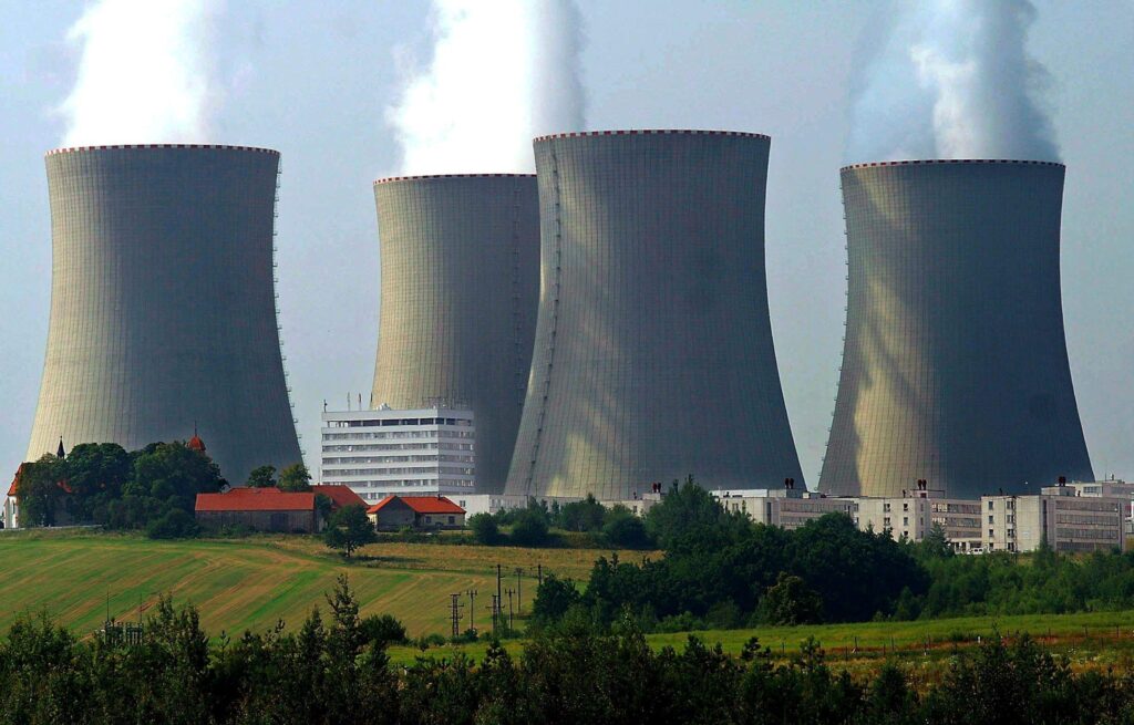 La imagen muestra una planta de energ?a nuclear con cuatro enormes torres de refrigeración liberando vapor, rodeada de edificios y un paisaje rural con campos y ?rboles.