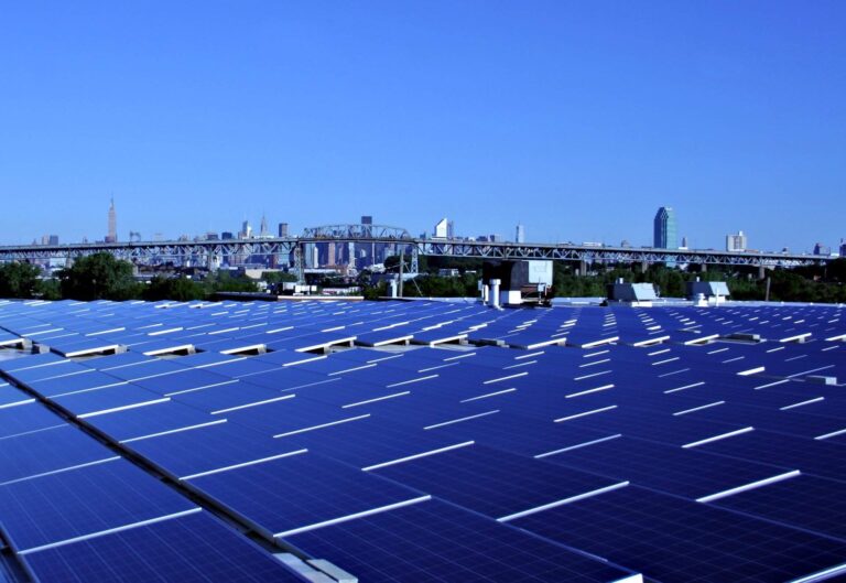 La imagen muestra una granja de paneles solares, con una ciudad en el fondo en el marco de un cielo azul sin nubes.