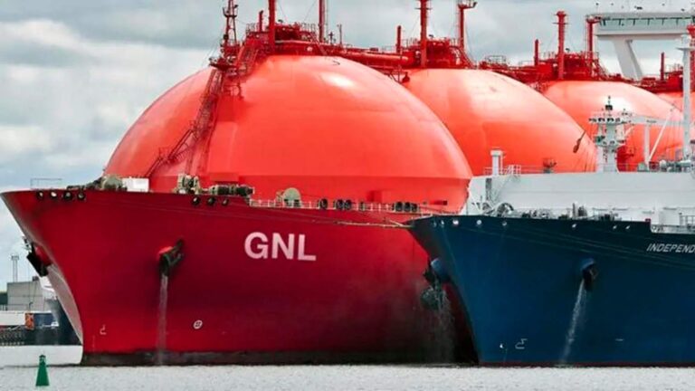 La imagen muestra buques especializados en el transporte de Gas Natural Licuado (GNL), con caracter?sticas distintivas como enormes tanques esféricos de color naranja, dise?ados para mantener el gas en estado l?quido a temperaturas extremadamente bajas. En el casco de uno de los buques se puede leer claramente "GNL". Estas embarcaciones se encuentran en lo que parece ser un puerto o ?rea de carga, con estructuras portuarias visibles al fondo.
