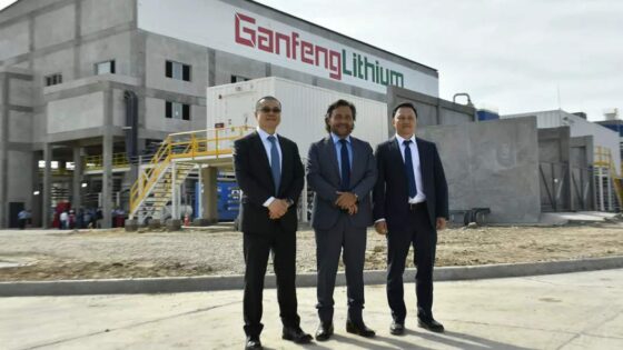 Tres hombres con traje y corbata posan frente a un edificio similar a una fábrica o planta con el logo de Ganfeng Lithium en rojo y verde.