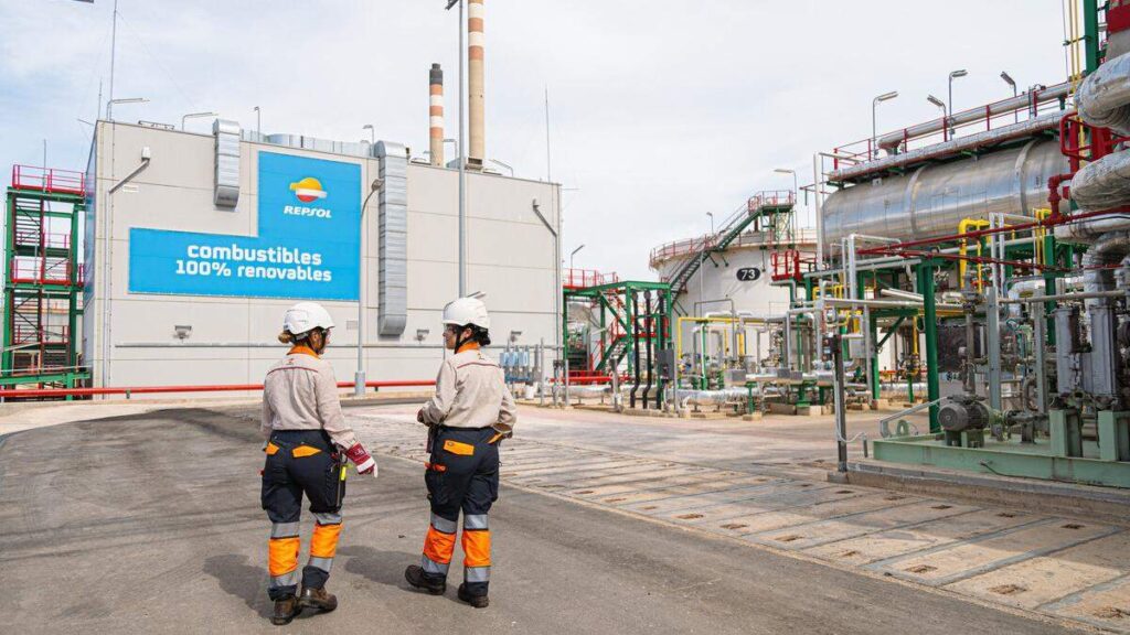 La imagen muestra a dos operarias con vestimenta de trabajo y casco caminando por una refiner?a. En uno de los edificios se ve el logo de Repsol con la leyenda '100% Renovables'.