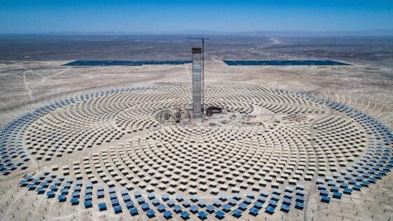 Vista aérea de la planta de energía termosolar Cerro Dominador en el desierto de Atacama, Chile. Se observa una torre central rodeada por un campo de heliostatos dispuestos en forma circular, y paneles solares planos en la distancia.