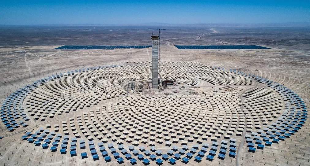 Vista aérea de la planta de energía termosolar Cerro Dominador en el desierto de Atacama, Chile. Se observa una torre central rodeada por un campo de heliostatos dispuestos en forma circular, y paneles solares planos en la distancia.