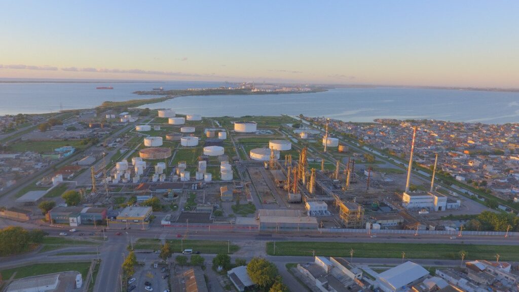 Vista aérea de una refiner?a de petróleo al amanecer, con tanques de almacenamiento blancos y estructuras industriales grises. La refiner?a se encuentra en una pen?nsula, rodeada de agua en tres lados y un ?rea urbana densamente poblada en el cuarto lado. Se ven carreteras y vegetación en primer plano, y un cuerpo de agua extenso con barcos en el horizonte.