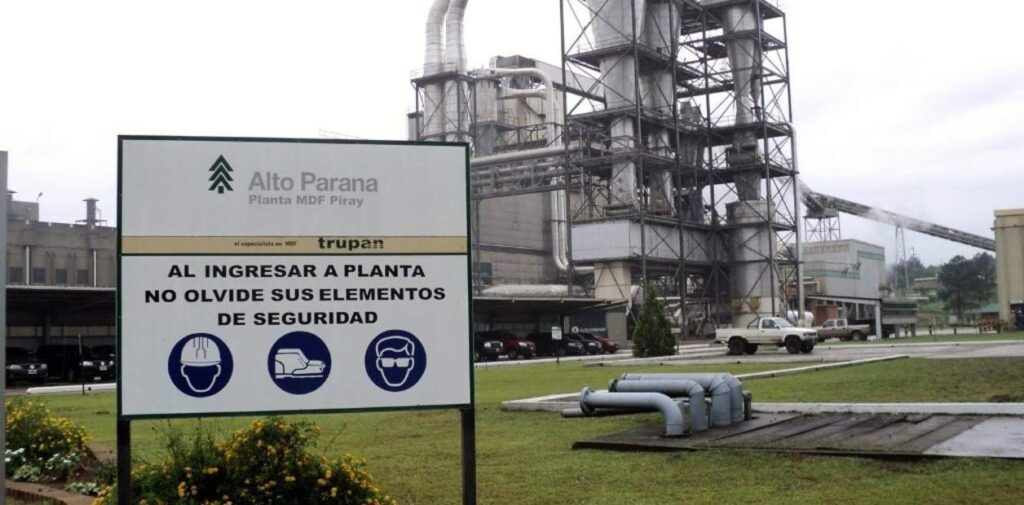 Planta industrial con letrero de seguridad en primer plano. Se ven estructuras de metal, tuberías y veh?culos al fondo.