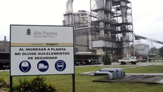Planta industrial con letrero de seguridad en primer plano. Se ven estructuras de metal, tuberías y vehículos al fondo.