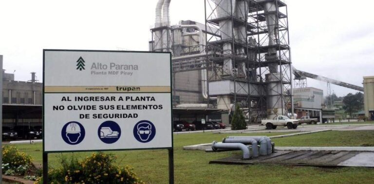 Planta industrial con letrero de seguridad en primer plano. Se ven estructuras de metal, tuberías y veh?culos al fondo.