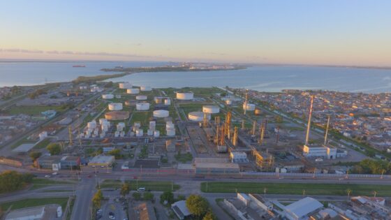 Vista aérea de una refinería de petróleo al amanecer, con tanques de almacenamiento blancos y estructuras industriales grises. La refinería se encuentra en una península, rodeada de agua en tres lados y un área urbana densamente poblada en el cuarto lado. Se ven carreteras y vegetación en primer plano, y un cuerpo de agua extenso con barcos en el horizonte.