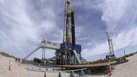 La imagen muestra una torre de perforación petrolera en un amplio terreno desértico. La torre se alza contra un cielo azul claro con nubes dispersas. En primer plano, se observa una plataforma de trabajo con barandas y escaleras, rodeada de conos de seguridad naranjas y una valla metálica. Al fondo, se distingue otra torre de perforación más pequeña.