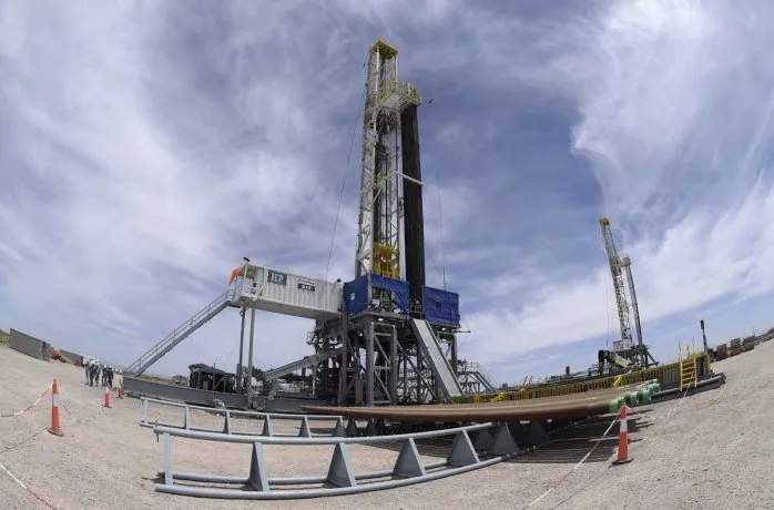 La imagen muestra una torre de perforación petrolera en un amplio terreno des?rtico. La torre se alza contra un cielo azul claro con nubes dispersas. En primer plano, se observa una plataforma de trabajo con barandas y escaleras, rodeada de conos de seguridad naranjas y una valla met?lica. Al fondo, se distingue otra torre de perforación m?s peque
