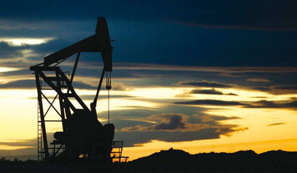 Silueta de una bomba extractora de petróleo (pumpjack) operando al atardecer en un campo petrolero, con un cielo dram?tico de nubes iluminadas por la luz del sol poniente.