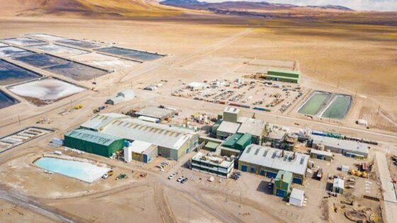 Vista aérea de una planta industrial en un paisaje árido con montañas al fondo. Se observan piscinas de evaporación de litio, edificios de procesamiento y maquinaria pesada.