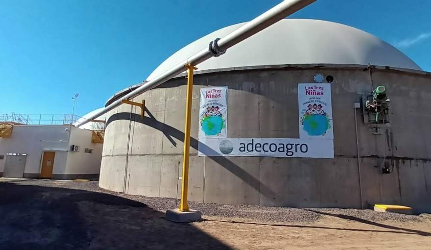 Imagen de un edificio industrial de gran tamaño con forma cil?ndrica y techo abovedado, con carteles de "Adecoagro" y "Las Tres Ni?as" en la fachada. Se observan tuberías met?licas amarillas y blancas que recorren el exterior del edificio. El cielo est? despejado y el sol ilumina la escena.
