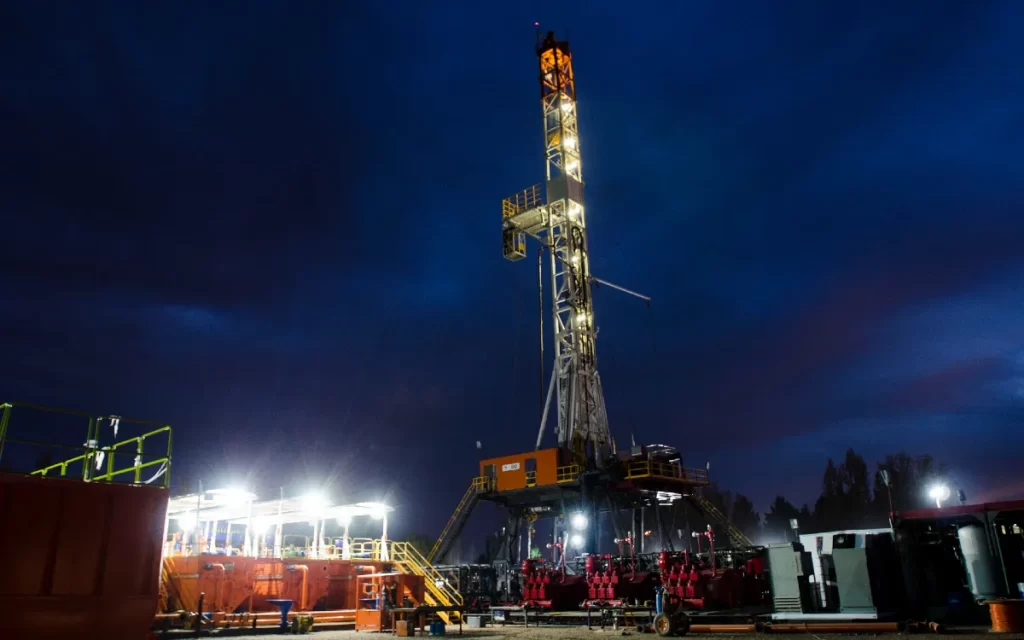Fotograf?a nocturna de una torre de perforación petrolera iluminada contra un cielo oscuro con tonos azules y naranjas del atardecer.