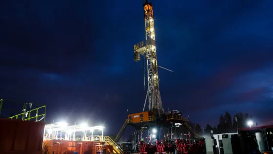 Fotografía nocturna de una torre de perforación petrolera iluminada contra un cielo oscuro con tonos azules y naranjas del atardecer.