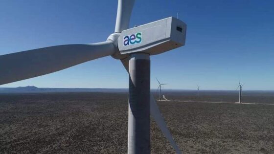 Primer plano de un aerogenerador con el logotipo de "aes" en su estructura, ubicado en un parque eólico en una zona árida y extensa. Al fondo, se observan más turbinas eólicas alineadas, bajo un cielo despejado y azul con un paisaje llano.