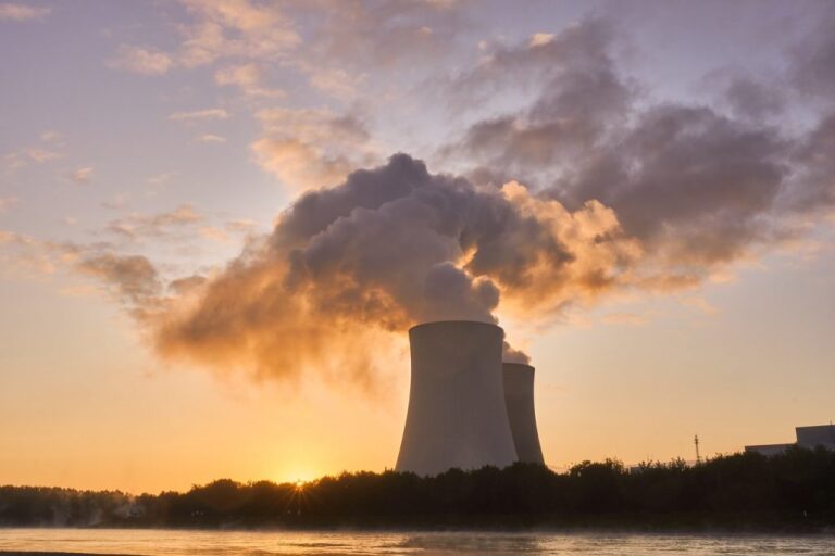 Vista al atardecer de una central nuclear con dos torres de refrigeración expulsando vapor de agua, iluminadas por la luz del sol poniente. La escena est? enmarcada por un cielo parcialmente nublado y una l?nea de ?rboles frente a un cuerpo de agua.