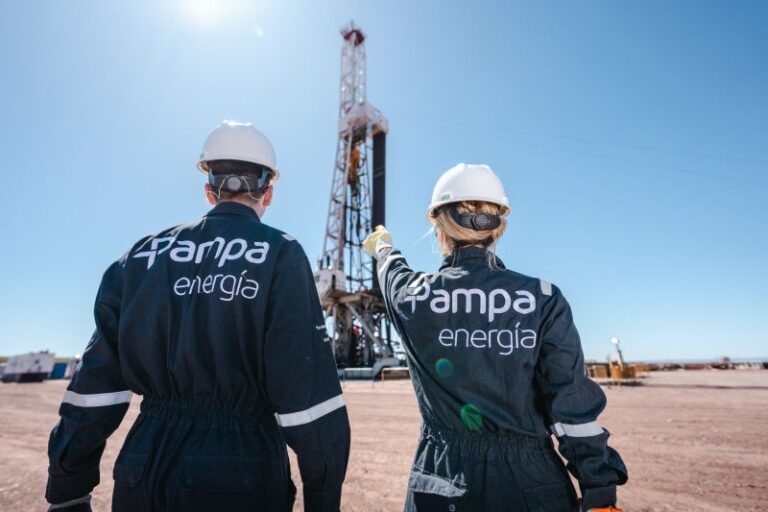 Dos trabajadores con cascos y overoles con el logo de "Pampa Energ?a" observan una torre de perforación petrolera en medio de un terreno ?rido, bajo un cielo despejado. Uno de ellos se?ala hacia la estructura.
