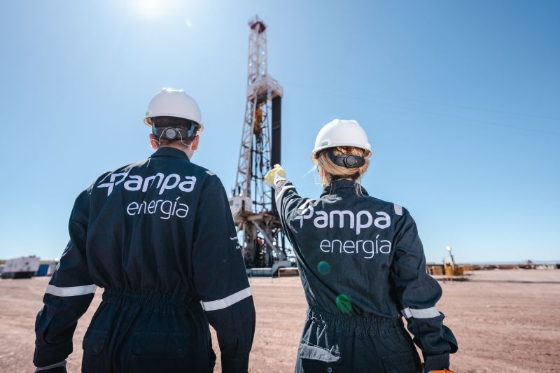 Dos trabajadores con cascos y overoles con el logo de "Pampa Energ?a" observan una torre de perforación petrolera en medio de un terreno ?rido, bajo un cielo despejado. Uno de ellos se?ala hacia la estructura.
