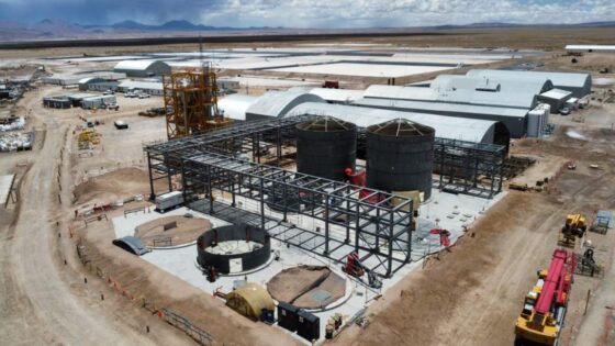 Vista aérea de una planta industrial en construcción en una zona desértica. El entorno muestra maquinaria pesada, caminos de tierra y un paisaje árido con montañas en el horizonte bajo un cielo nublado.