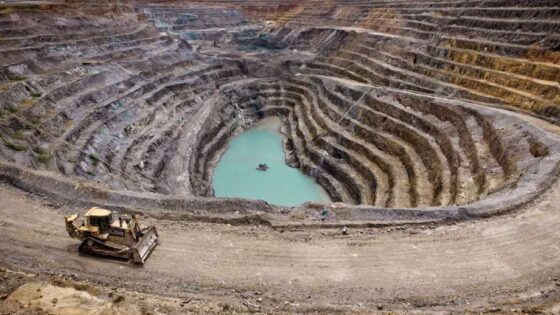 Vista aérea de una mina a cielo abierto con terrazas concéntricas, una excavadora en primer plano y un pequeño cuerpo de agua turquesa en el fondo del cráter.