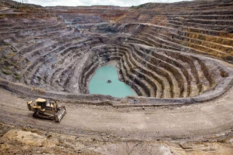 Vista aérea de una mina a cielo abierto con terrazas conc?ntricas, una excavadora en primer plano y un peque?o cuerpo de agua turquesa en el fondo del cr?ter.