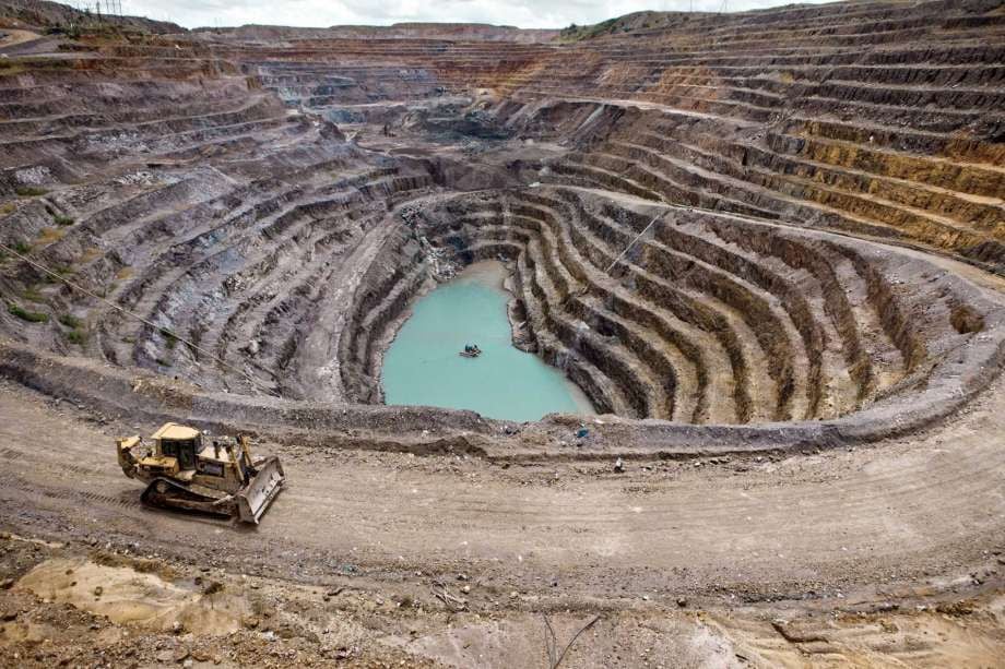 Vista aérea de una mina a cielo abierto con terrazas concéntricas, una excavadora en primer plano y un pequeño cuerpo de agua turquesa en el fondo del cráter.