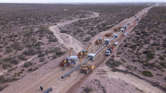 Vista aérea de una línea de construcción de gasoductos extendiéndose a través de un terreno árido y seco. Varias máquinas de gran tamaño y vehículos de trabajo, junto con personal, están posicionados a lo largo.