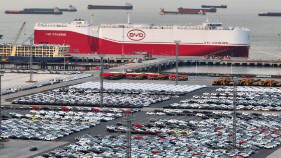 Buque portavehículos de BYD cargando autos eléctricos en un puerto chino, con miles de unidades listas para exportación. Al fondo, se destacan embarcaciones de carga y el letrero “LNG Powered”.