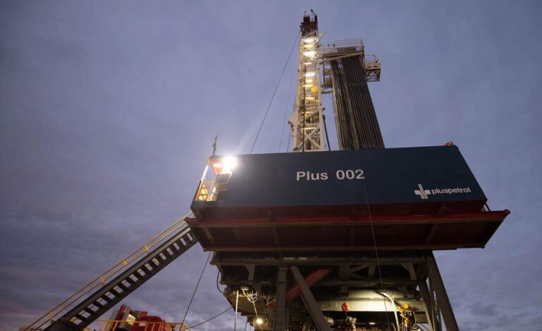 Torre de perforación petrolera de Pluspetrol, iluminada al anochecer, con estructura metálica y trabajadores operando en la base.