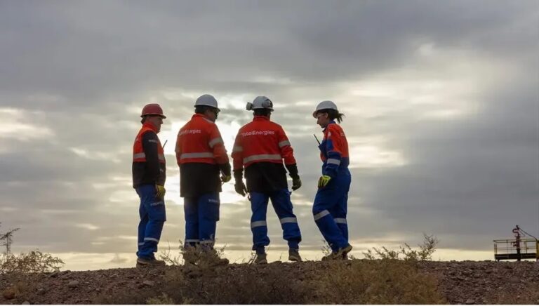 Cuatro personas con cascos y uniformes de seguridad de TotalEnergies (chaquetas rojas con el logo, pantalones azules y cintas reflectivas) conversan al aire libre, sobre un terreno ?rido. El cielo est? nublado y crea una atm?sfera de contraluz que resalta las siluetas de los trabajadores. Al fondo, se distingue una estructura industrial.