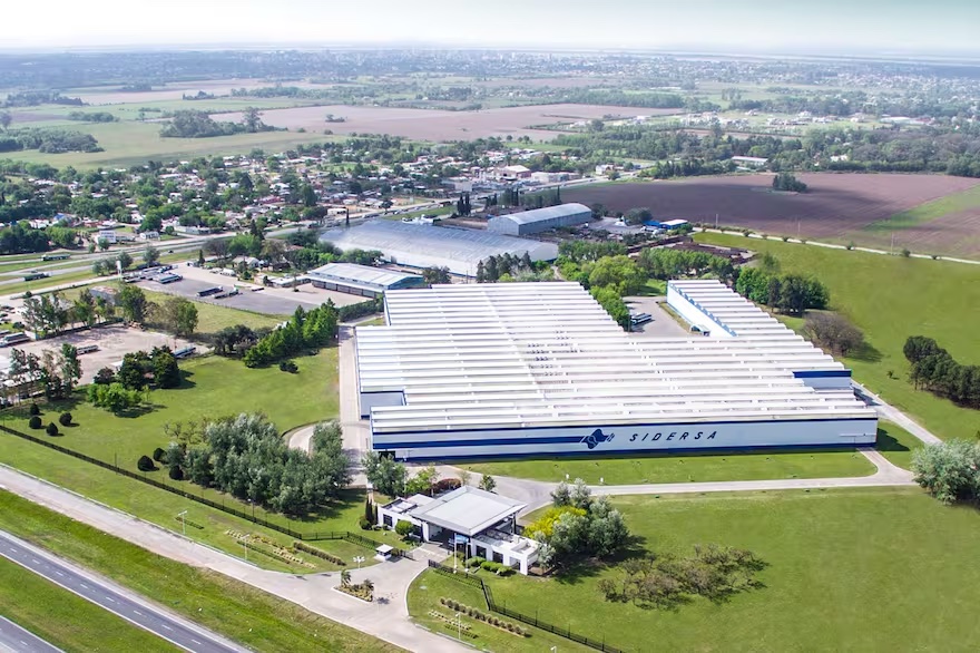 Vista aérea de la planta industrial de Sidersa en San Nicol?s. Se observan grandes estructuras met?licas, dep?sitos, chimeneas y ?reas de carga, todo rodeado por vegetación y caminos internos. El cielo est? despejado y se aprecia parte del entorno urbano e industrial cercano.
