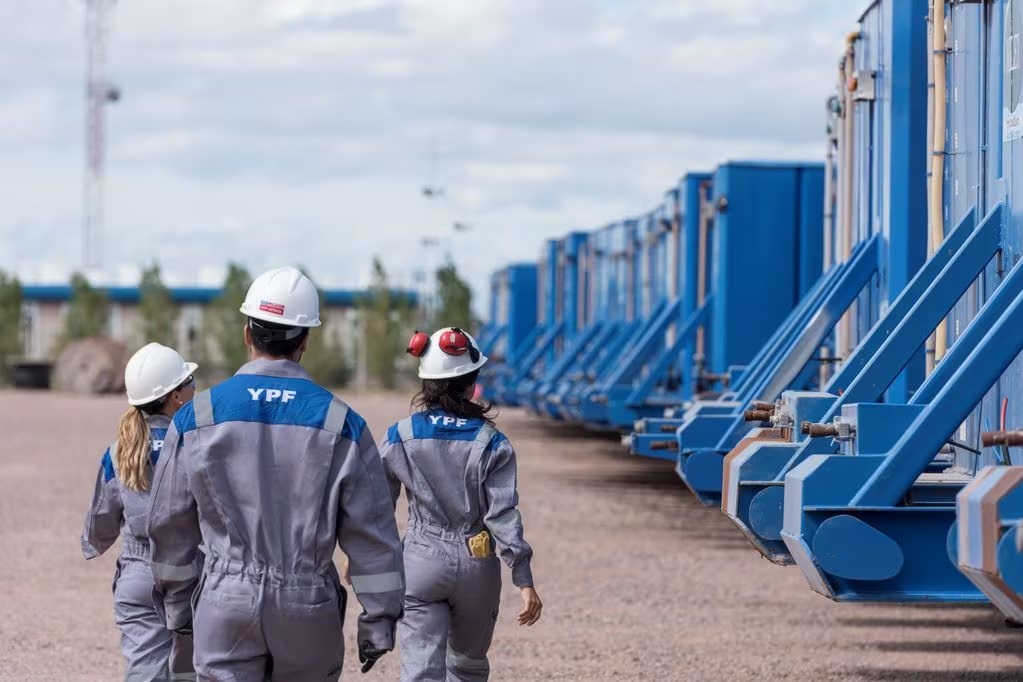 Tres trabajadores de YPF, con mamelucos grises y cascos de seguridad, caminan por una planta industrial al aire libre. A su derecha, se observa una larga fila de estructuras azules alineadas, posiblemente contenedores o equipos t?cnicos. El cielo est? parcialmente nublado y el entorno es amplio y despejado.