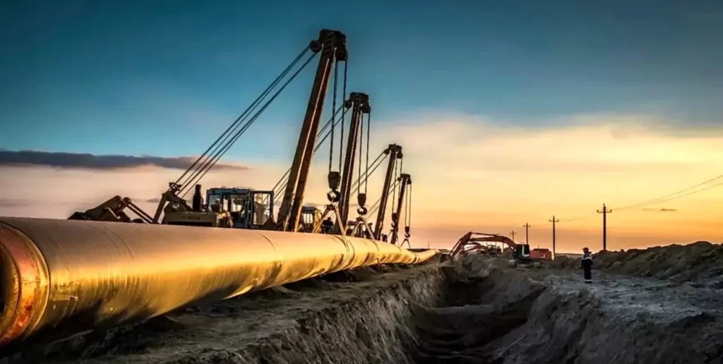 Obras de instalación de un gasoducto al atardecer, con maquinaria pesada levantando tuberías y trabajadores en el sitio de construcci