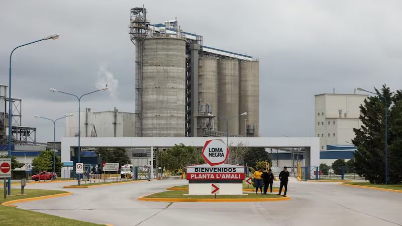 Entrada principal de la planta L?Amal? de Loma Negra, con silos industriales al fondo y personas caminando por el acceso.