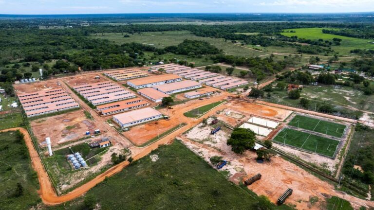 Vista aérea de un complejo industrial en construcción rodeado de áreas verdes y caminos de tierra, con canchas deportivas visibles hacia la derecha.