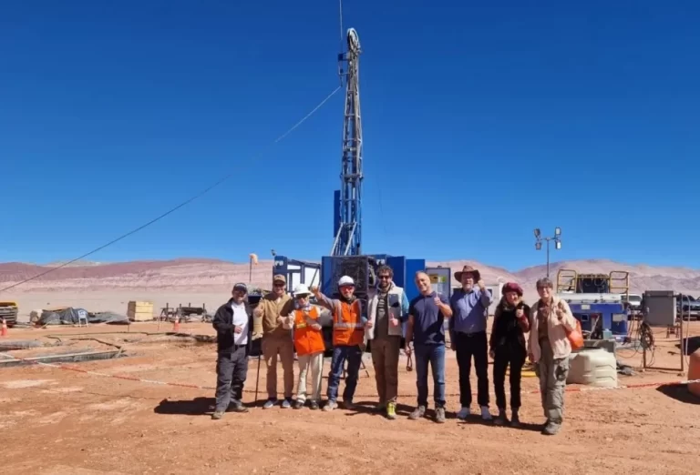Grupo de personas con indumentaria de trabajo posando frente a una torre de perforación en una zona desértica bajo un cielo despejado.