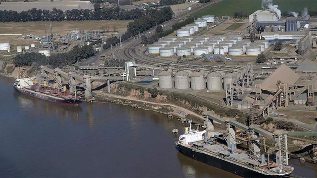 Vista aérea del complejo portuario e industrial de Timb?es, con buques de carga amarrados sobre el r?o Paran? y estructuras de silos, cintas transportadoras y tanques de almacenamiento en tierra.