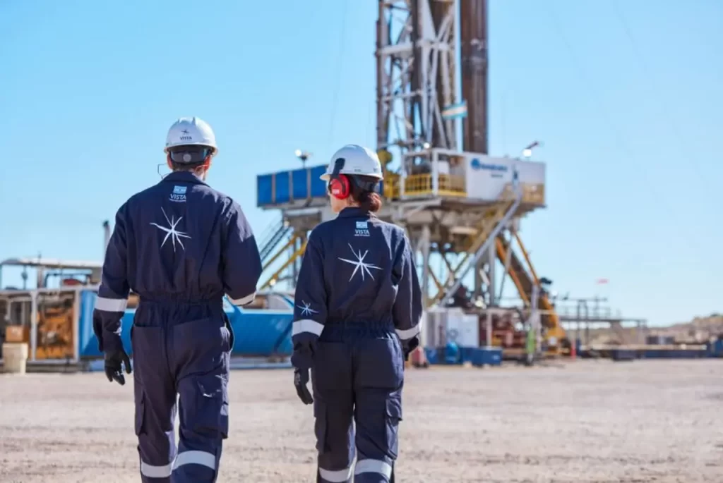Dos trabajadores con uniformes de "VISTA" y cascos de seguridad caminan hacia una torre de perforación en un sitio industrial.