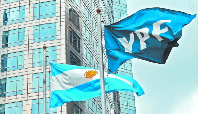 Banderas de YPF y de Argentina ondeando frente a un edificio de oficinas de vidrio.