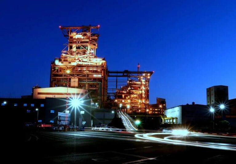 Vista nocturna de una planta industrial iluminada, con una estructura metálica de gran altura y luces anaranjadas, mientras vehículos dejan estelas de luz sobre la carretera en primer plano.