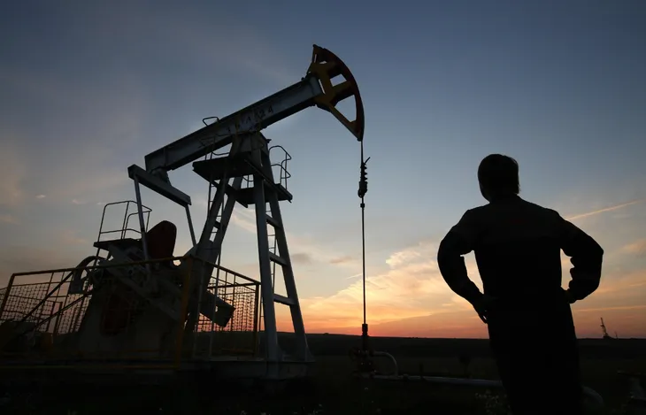 Silueta de un trabajador frente a un equipo de bombeo petrolero (pumpjack) en operación, con el cielo al atardecer de fondo.