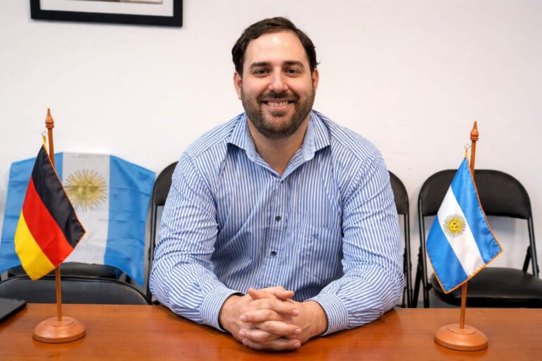 Hombre sonriente sentado detrás de una mesa, con las manos entrelazadas, flanqueado por pequeñas banderas de Argentina y Alemania en un entorno institucional.