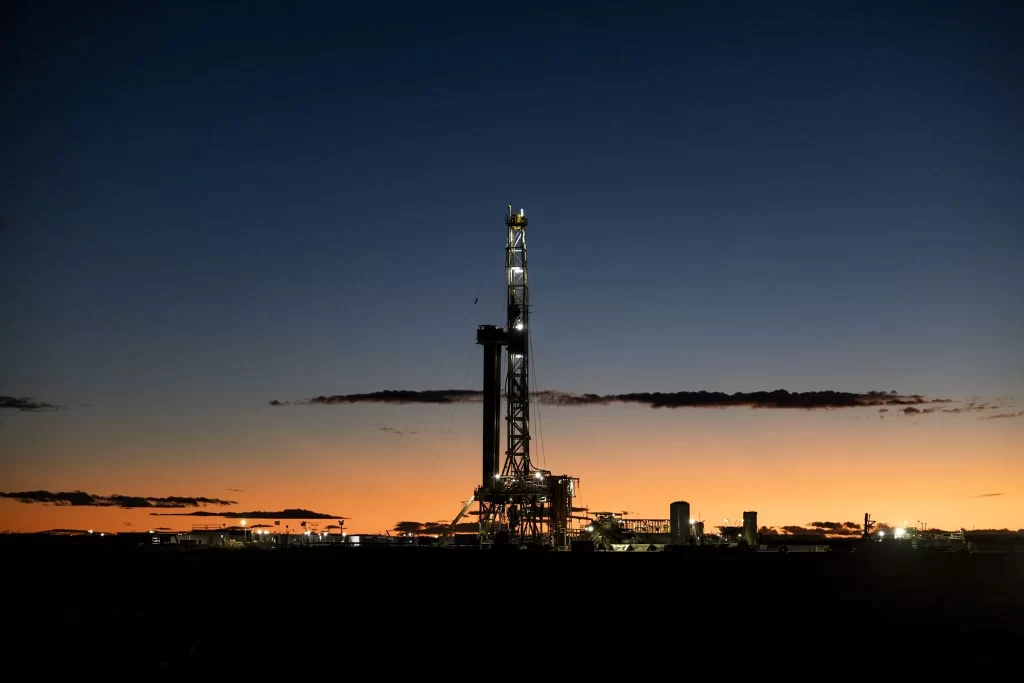 Torre de perforación petrolera iluminada en Vaca Muerta durante el atardecer, recortada en silueta contra un cielo azul y naranja, con instalaciones industriales visibles en el horizonte.
