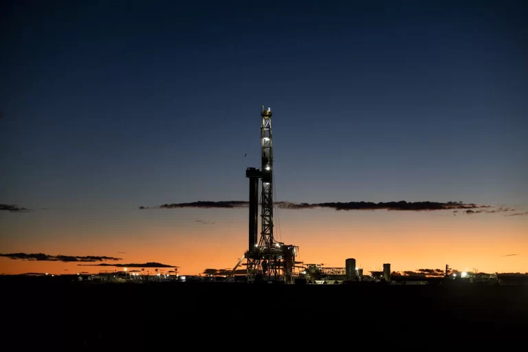 Torre de perforación petrolera iluminada en Vaca Muerta durante el atardecer, recortada en silueta contra un cielo azul y naranja, con instalaciones industriales visibles en el horizonte.