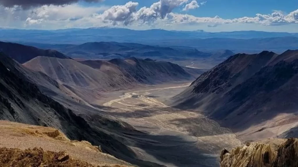 Vista panorámica de un valle montañoso con río serpenteante en el fondo, rodeado de altas laderas áridas bajo cielo parcialmente nublado.