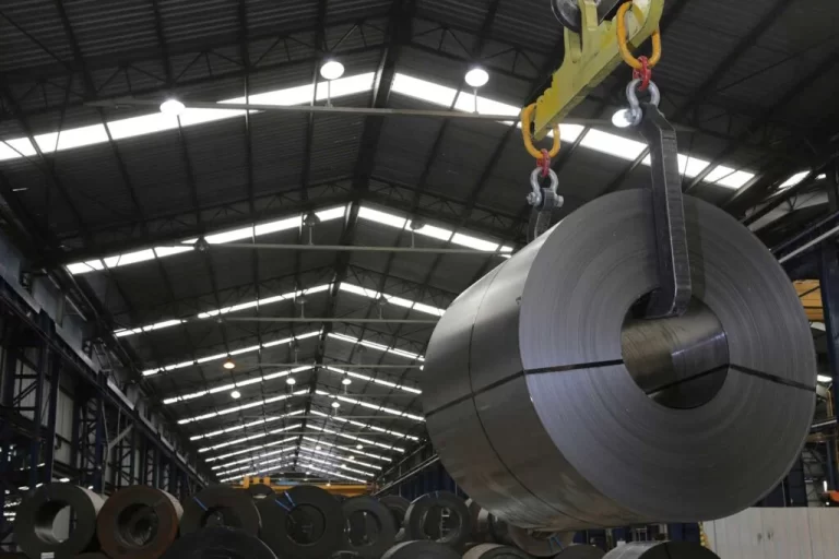 Bobina de acero suspendida por una grúa puente dentro de una planta sider?rgica, con otras bobinas met?licas apiladas en el interior de una nave industrial.